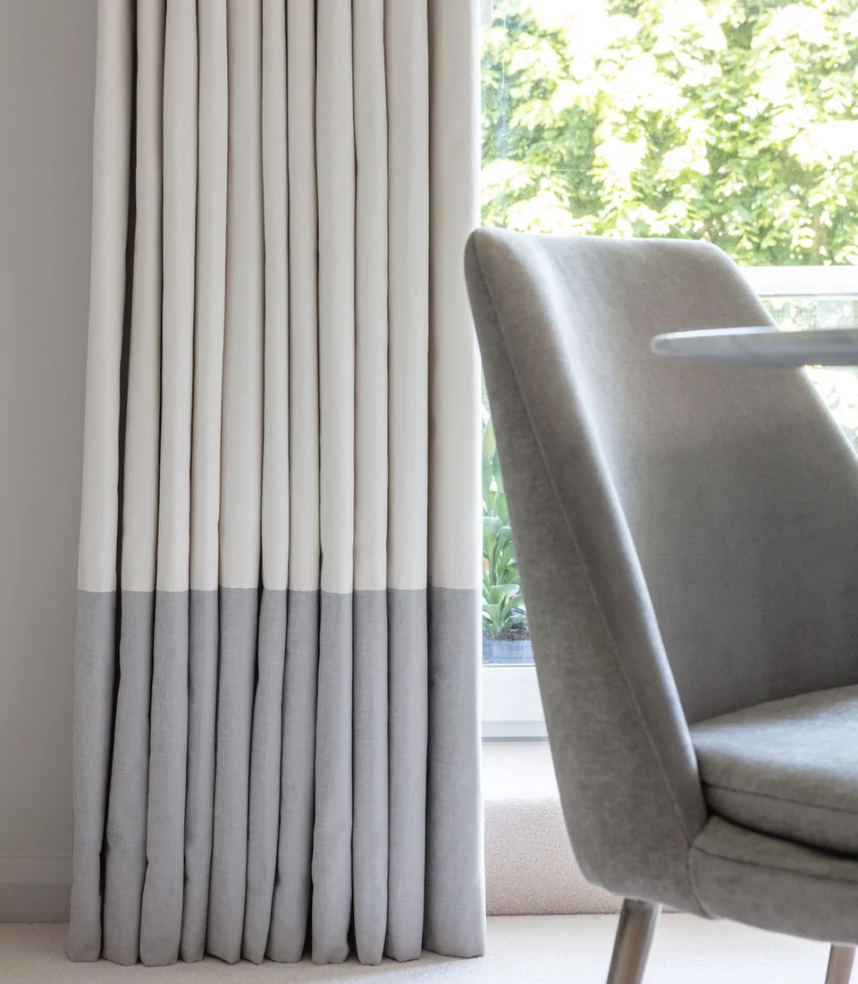 A pair of neatly folded curtains with a light cream top and gray bottom hangs beside a modern gray chair, framed by a bright window overlooking greenery outside.