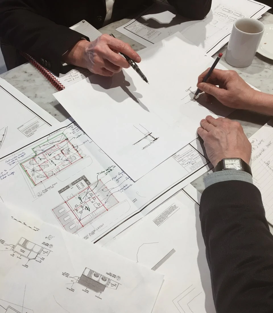 Two hands are engaged in sketching architectural plans on papers spread across a table, surrounded by notebooks, a cup, and a plate, indicating collaboration in a design discussion.