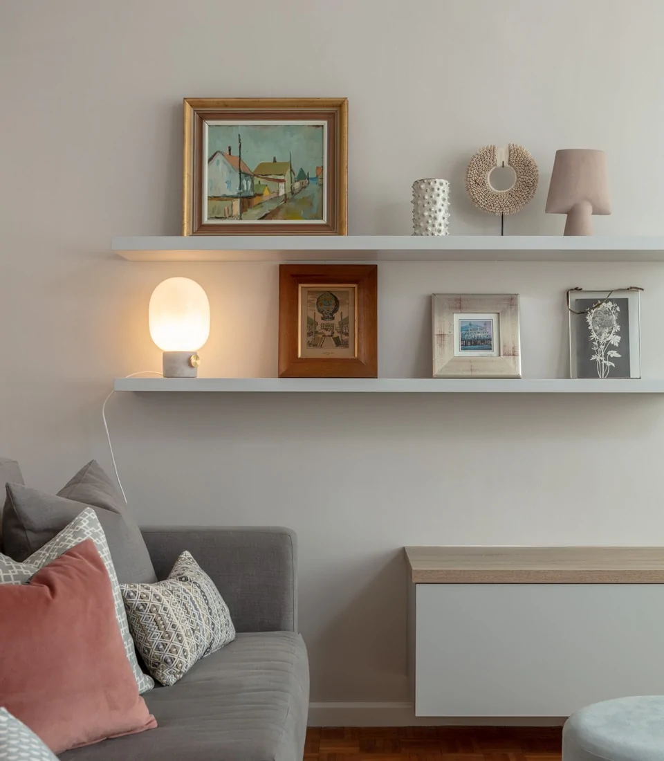 A modern living space features a gray sofa adorned with decorative pillows. Above, a white shelf holds paintings and decorative objects, including a lamp casting a warm glow.