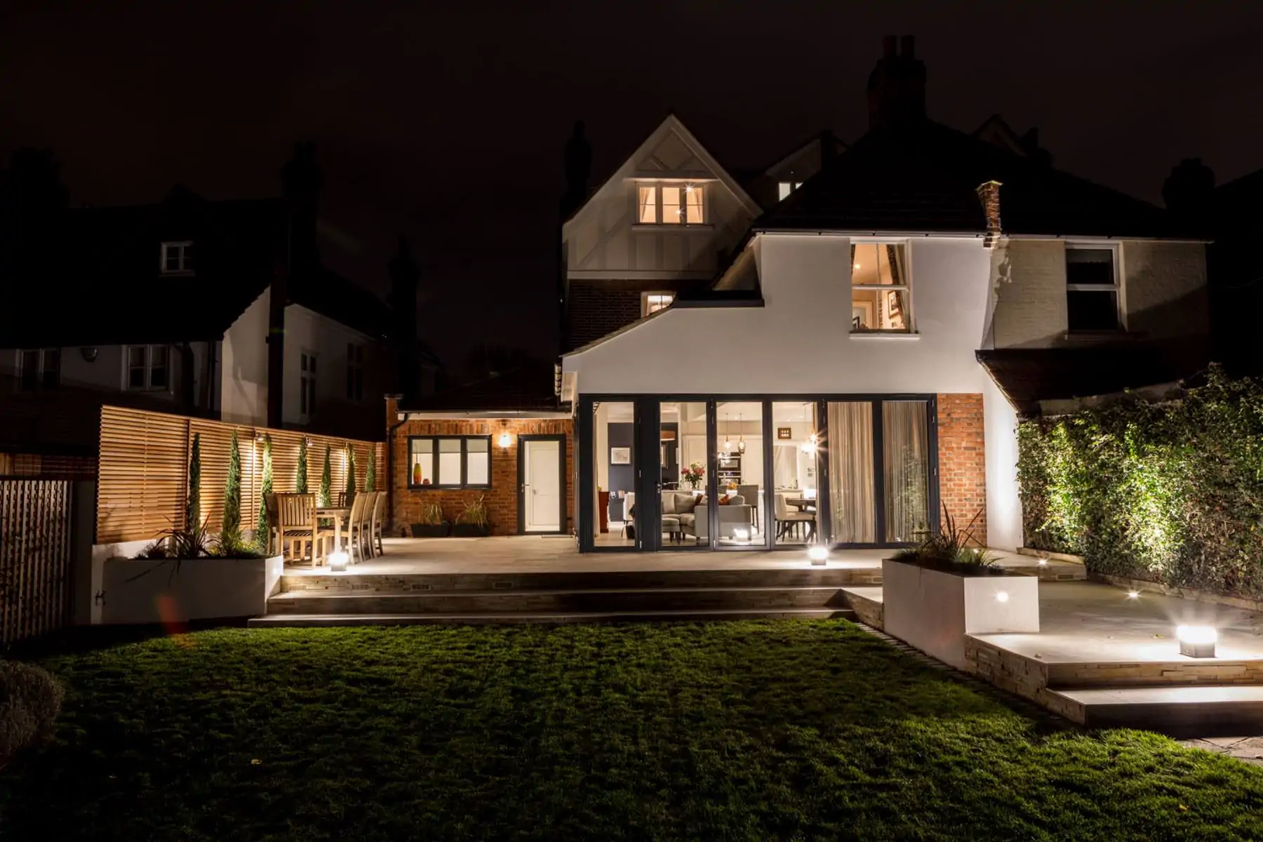 A well-lit modern home features a patio with outdoor furniture and greenery, illuminated by soft lights, surrounded by dark skies and neighboring houses at night.