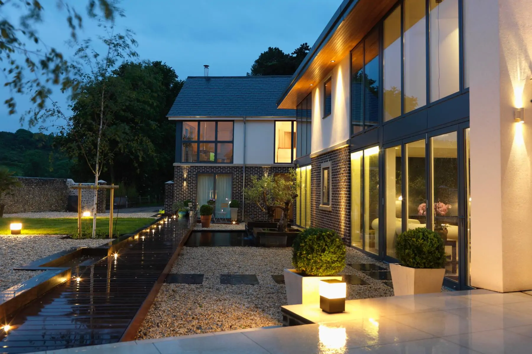 A modern house features large glass windows and a wooden deck illuminated by soft lights. Surrounded by a landscaped garden, the setting is serene during twilight.