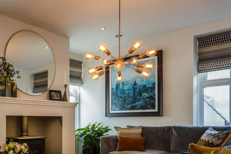 A modern chandelier with multiple exposed bulbs hangs from the ceiling. Below, a cozy living room features a gray sofa with decorative cushions, a round mirror, and a large artwork on the wall.