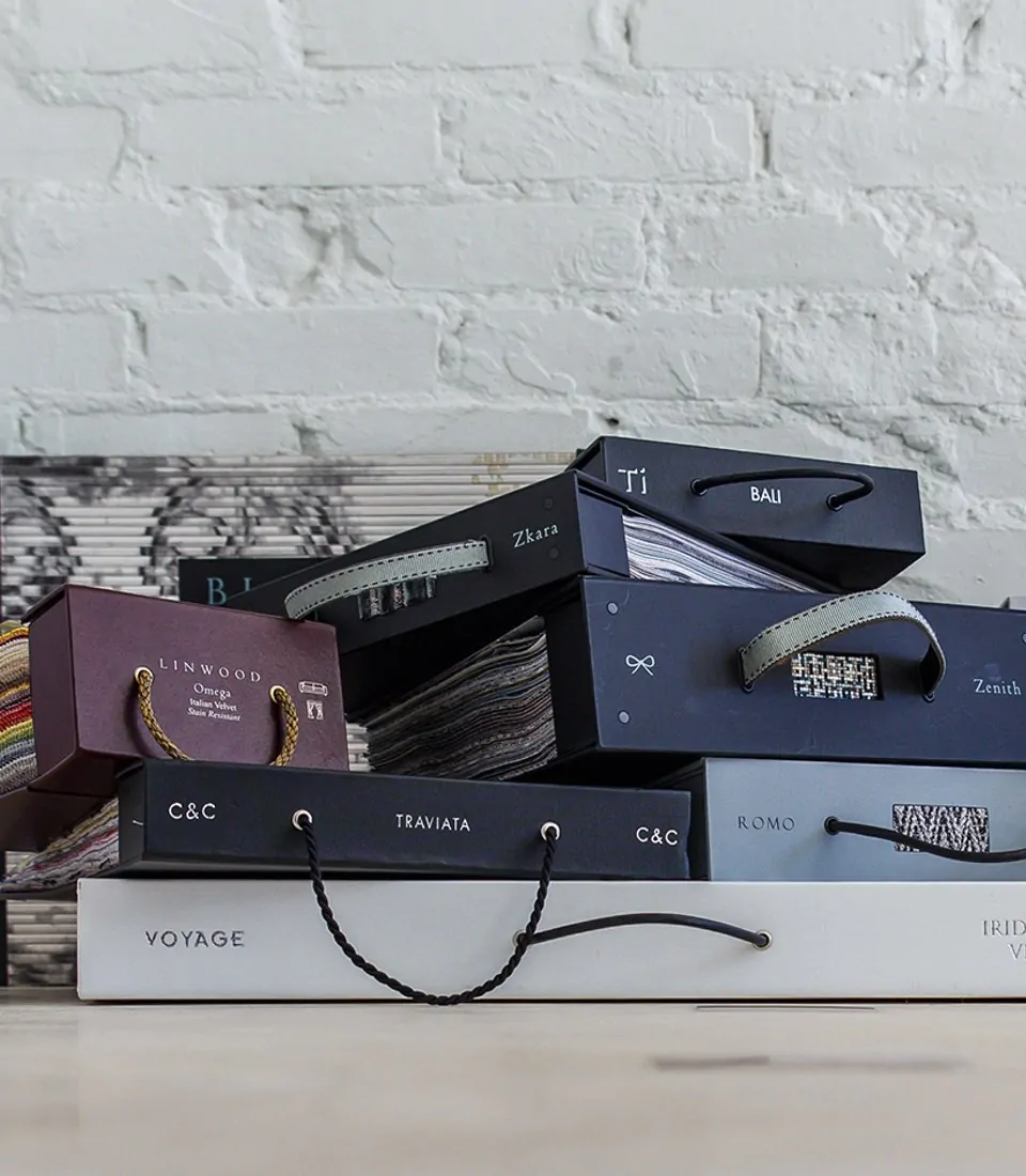 A collection of stacked sample boxes with handles, showcasing various designs and colors. The background features textured white brick, enhancing the aesthetic of the neatly arranged objects.