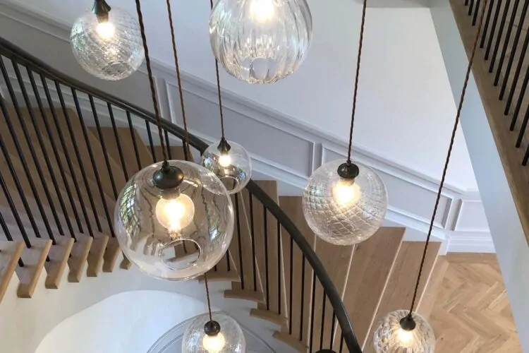 Hanging glass light fixtures illuminate a spiral staircase, casting soft light in a modern, minimalist interior. The stair treads are wooden, contrasted by a dark railing and white walls.