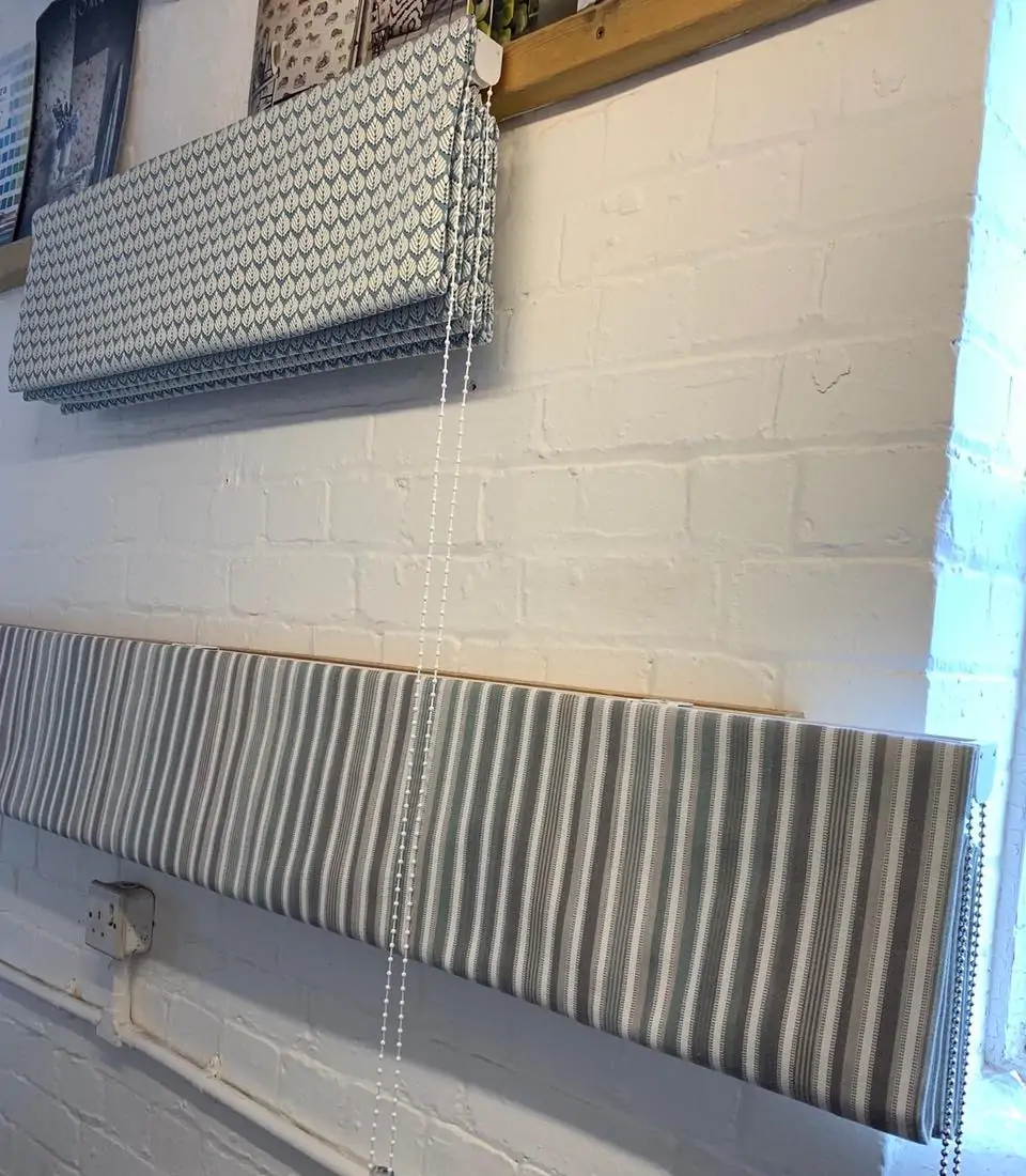 Roller blinds with leaf and striped patterns are displayed on a white brick wall. One blind is rolled up, while another is partially rolled down, showing the fabric designs. A window is visible in the background.