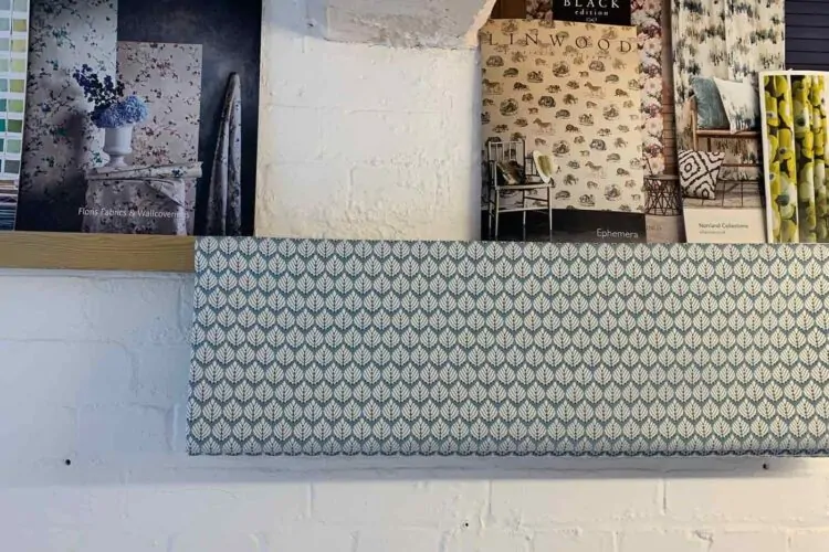 A shelf displays several fabric sample books, showcasing various floral and patterned designs, with a patterned fabric swatch hanging below, all set against a white brick wall.