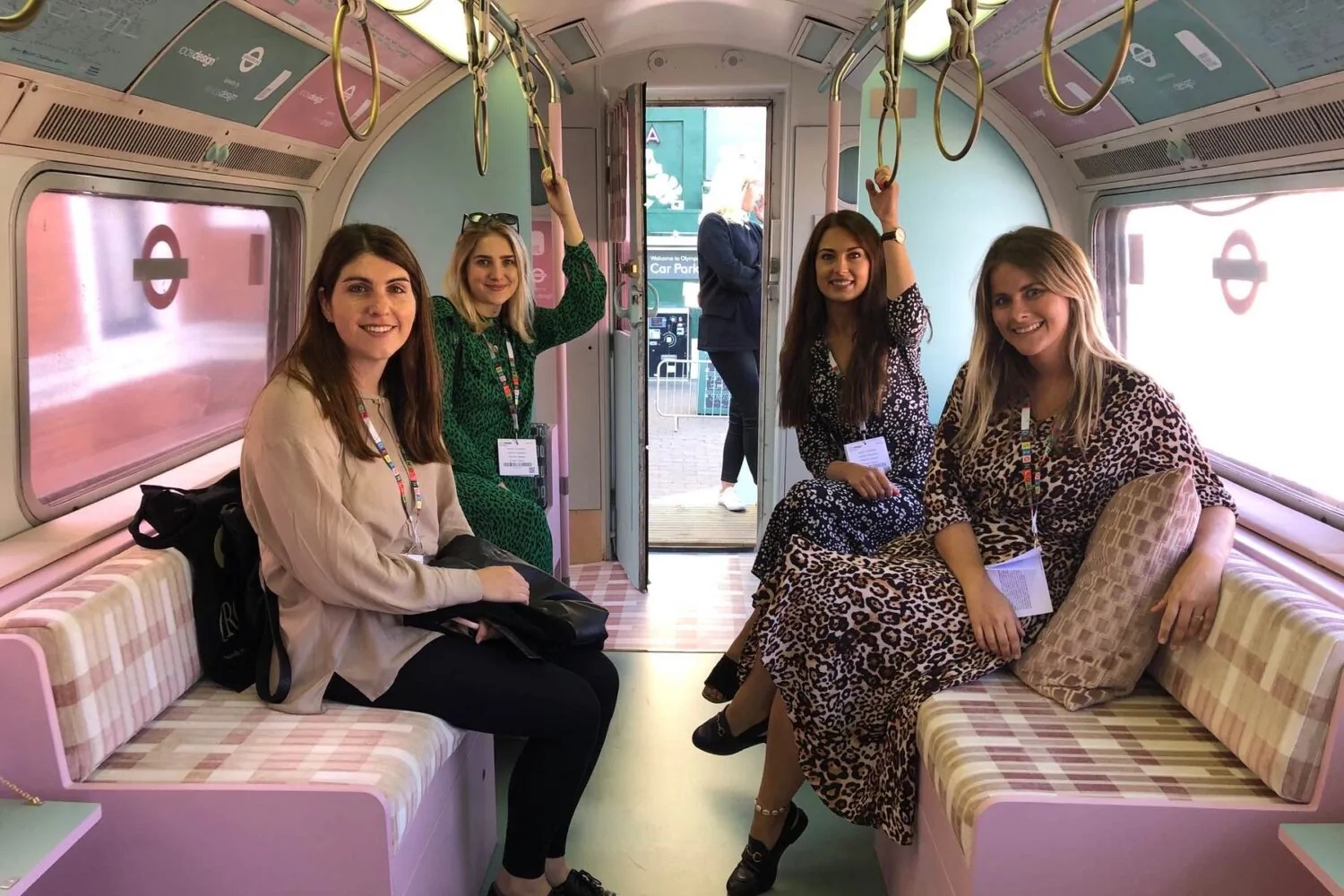 Four women are seated in a colorful, vintage-style train interior, smiling and posing for a photo. They are dressed in varied patterns, with a view of an open train door behind them.