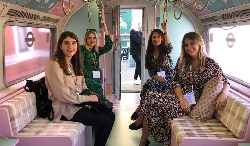 Four women are seated in a colorful, vintage-style train interior, smiling and posing for a photo. They are dressed in varied patterns, with a view of an open train door behind them.