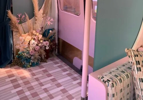 A floral arrangement in soft hues is positioned on a patterned floor near a pink and green wall. A cozy, patterned couch and curtains add a warm atmosphere to the inviting space.