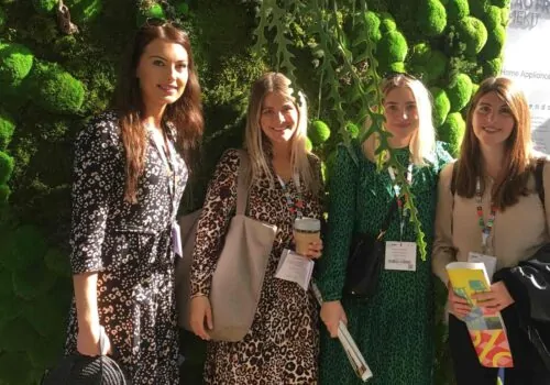 Four women stand together in front of a vibrant green moss wall. Each wears a stylish dress, and one holds a coffee cup, with various event badges around their necks.