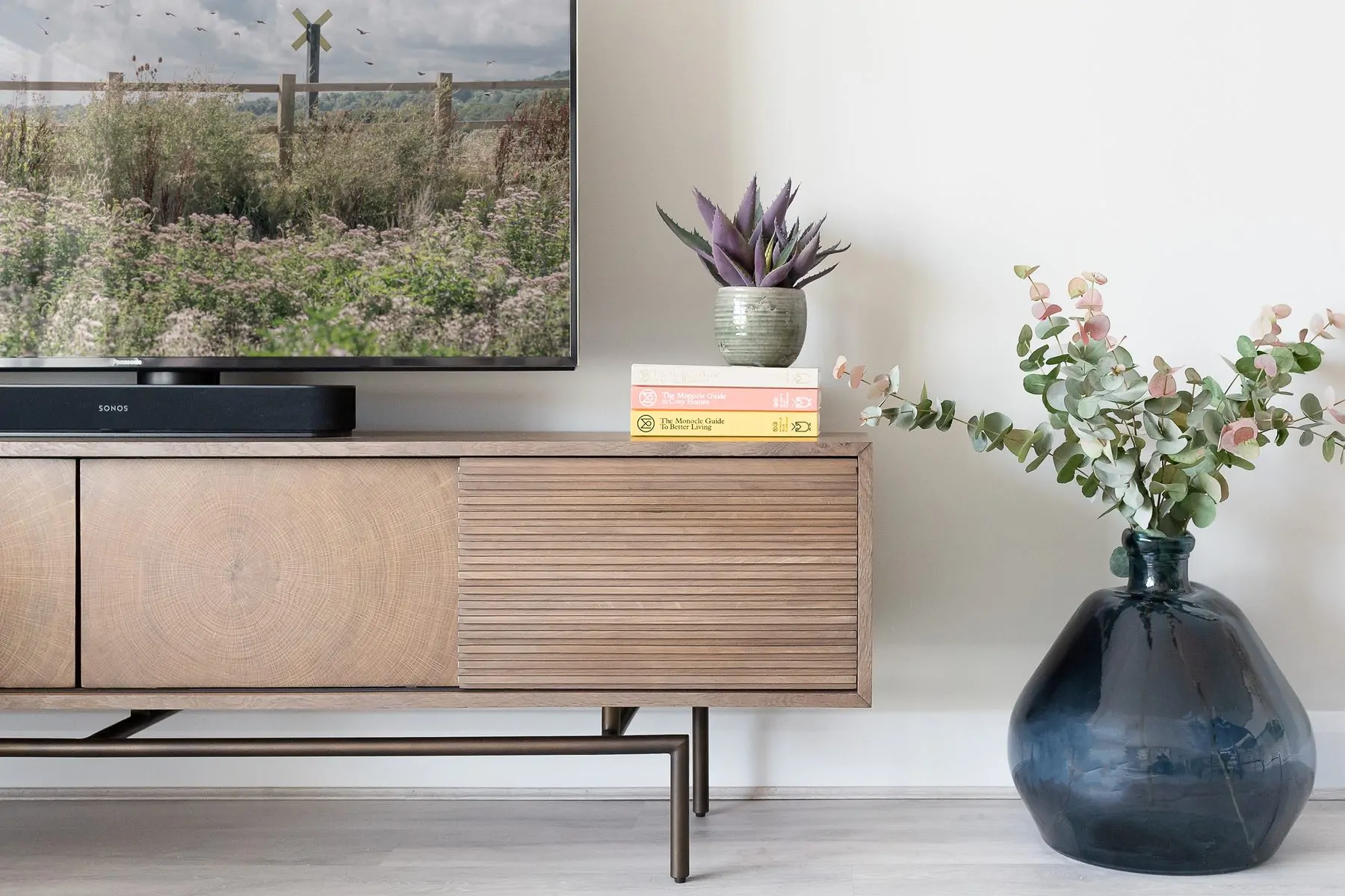 A wooden media console displays a television showing a scenic view. A speaker and stacked books rest atop. Potted plants and a large vase with greenery adorn the side.