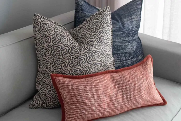 Three decorative pillows in various textures and colors rest on a light gray sofa. The background features soft fabric drapes, enhancing the cozy living space ambiance.
