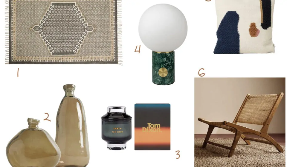 A decorative rug lies flat, two glass vases rest beside a candle in a black jar, a round lamp stands on a green marble base, and a textured throw pillow sits near a wooden chair on a woven mat.