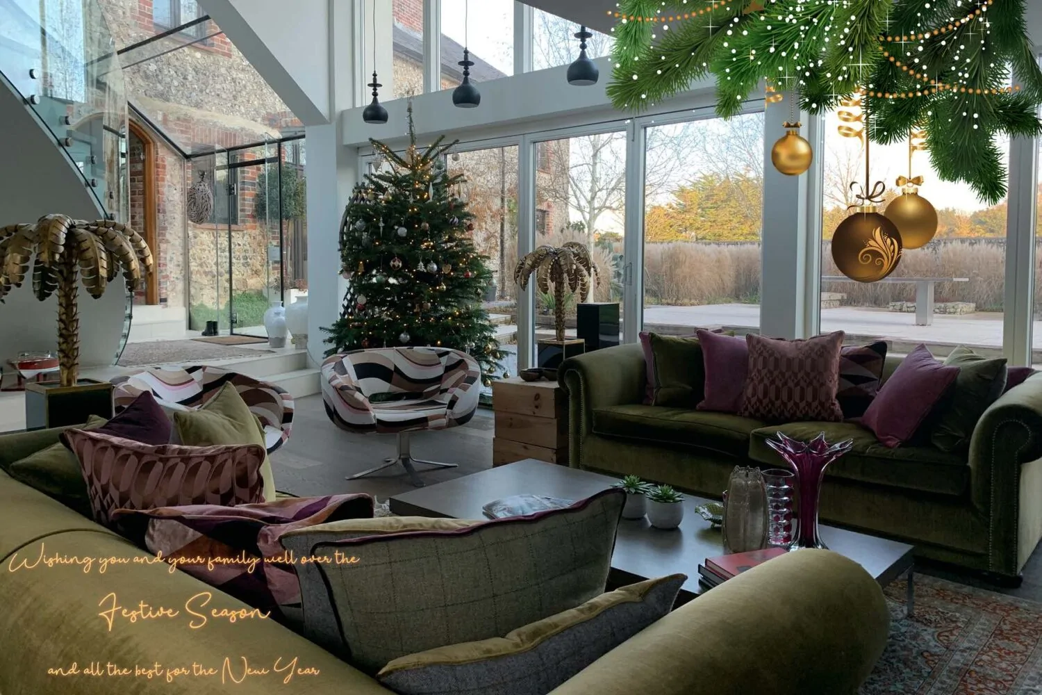 A beautifully decorated living room features a Christmas tree and a cozy seating area. Large glass windows display a scenic outdoor view, while hanging ornaments add a festive touch.