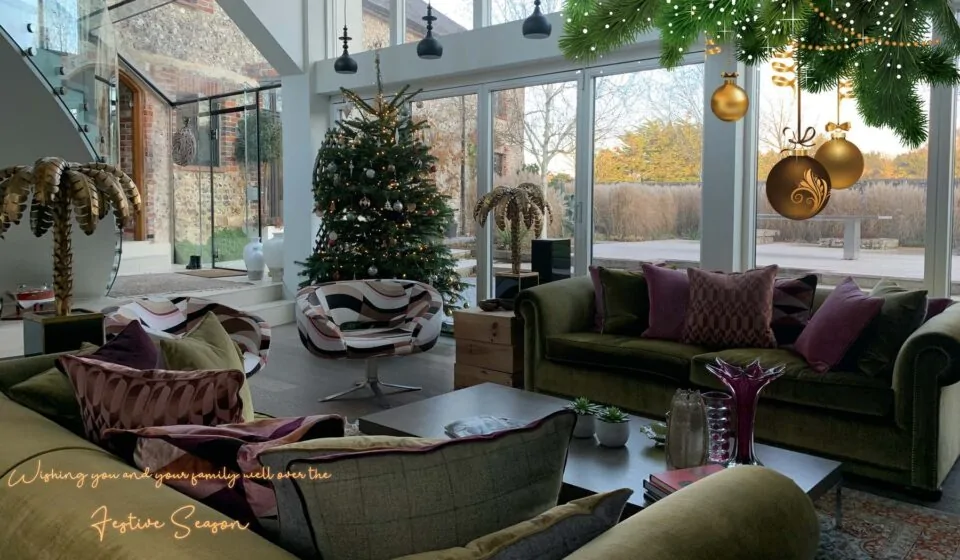 A beautifully decorated living room features a Christmas tree and a cozy seating area. Large glass windows display a scenic outdoor view, while hanging ornaments add a festive touch.