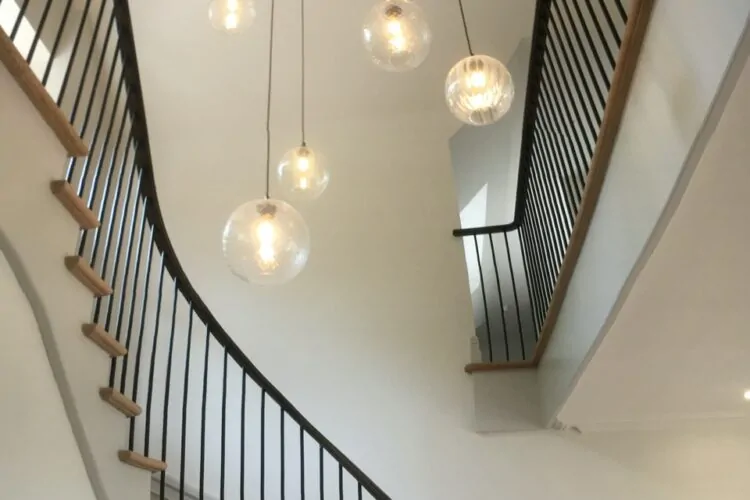 A curved staircase with wooden steps and black railings ascends beside a smooth white wall, illuminated by multiple hanging glass pendant lights.