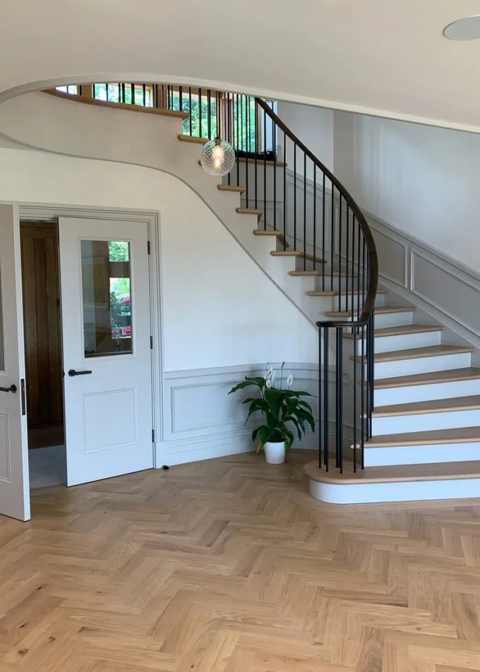 A curved staircase with wooden steps ascends to a landing in a well-lit foyer. Two white doors open to an adjacent room, while a potted plant adds greenery to the space.