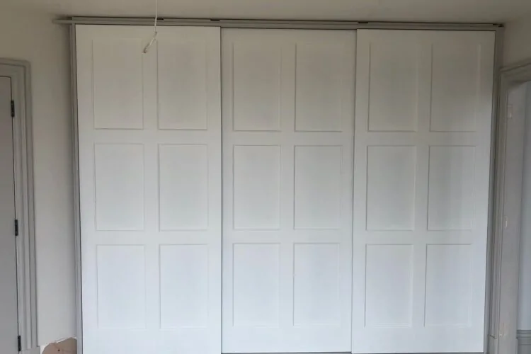 A set of three white, paneled sliding doors is positioned against a neutral wall. The surrounding area shows unfinished flooring and a partially framed doorway on the right.
