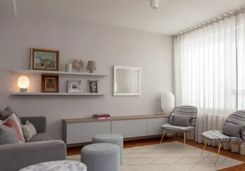A modern living room features a gray sofa with decorative pillows, two patterned chairs, circular tables, and a low cabinet. Shelves display framed art and a lamp, while sunlight filters through sheer curtains.