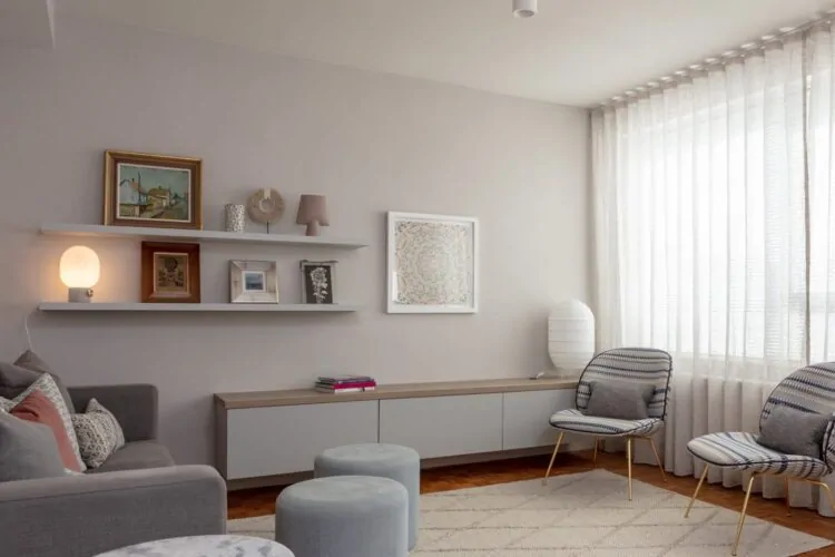 A modern living room features a gray sofa with decorative pillows, two patterned chairs, circular tables, and a low cabinet. Shelves display framed art and a lamp, while sunlight filters through sheer curtains.