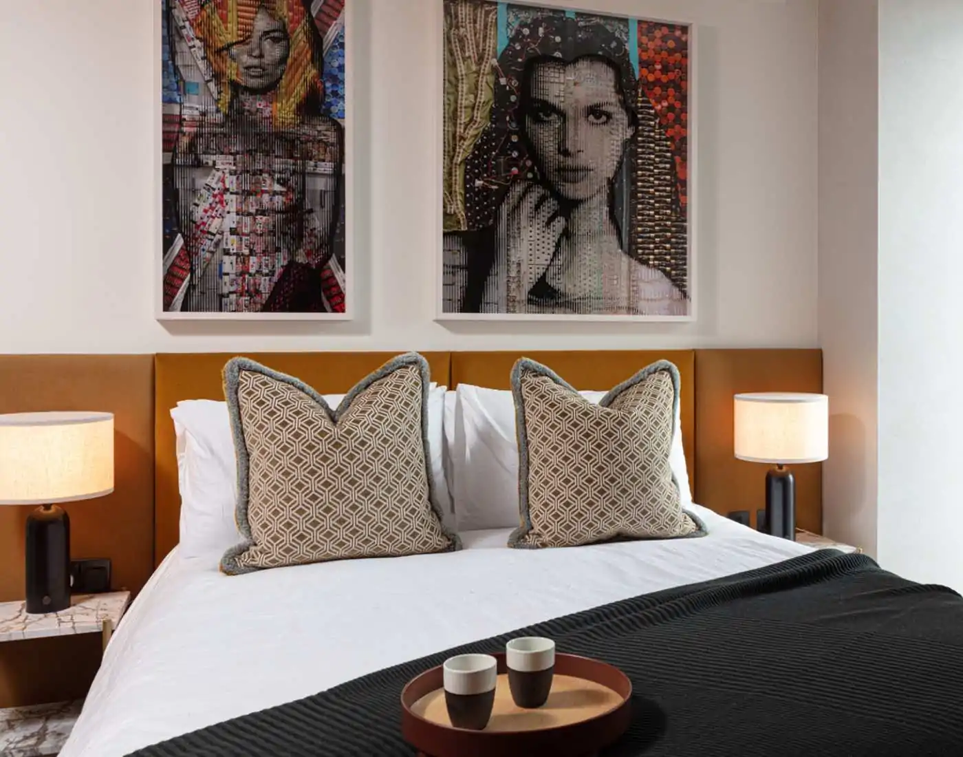 A stylish bed with decorative pillows is centered in a modern room. Above, two framed art pieces add color. A side table holds a tray with cups, complemented by ambient lamps.