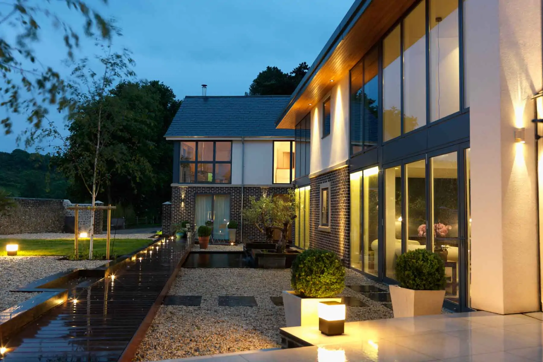 A modern house with large windows reflects warm lights in the evening. A wooden pathway leads through a landscaped garden with pebbles, planters, and subtle illumination, surrounded by trees and greenery.