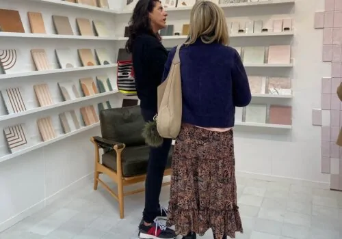 Two women are engaged in conversation in a gallery-like space, surrounded by shelves displaying various art pieces or notebooks, with a low wooden chair nearby.