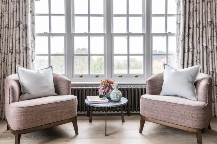 Two comfortable, modern armchairs face a small circular table topped with a vase and books. Natural light streams through large windows, revealing greenery outside, while elegant curtains frame the scene.