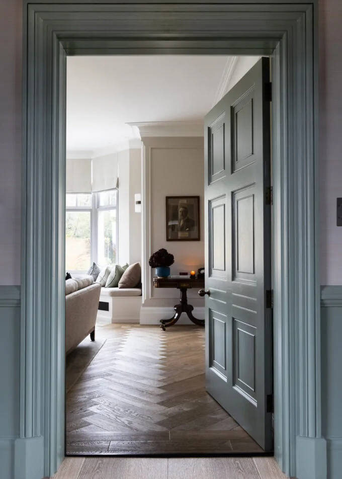 A partially open green door reveals a cozy room with a beige sofa, decorative pillows, and an elegant table with a candle, amid a lightly textured wooden floor.