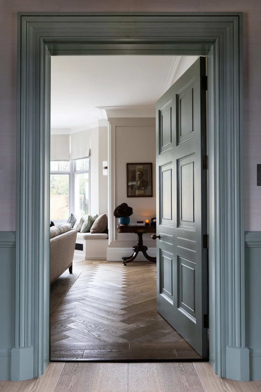 A partially open green door reveals a cozy room with a beige sofa, decorative pillows, and an elegant table with a candle, amid a lightly textured wooden floor.