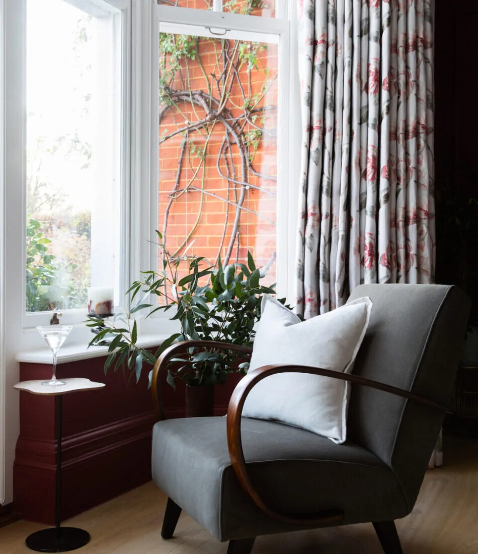 A modern gray chair with wooden arms sits beside a small table holding a martini glass. Nearby, a potted plant thrives in the sunny corner by large windows framed by floral curtains.