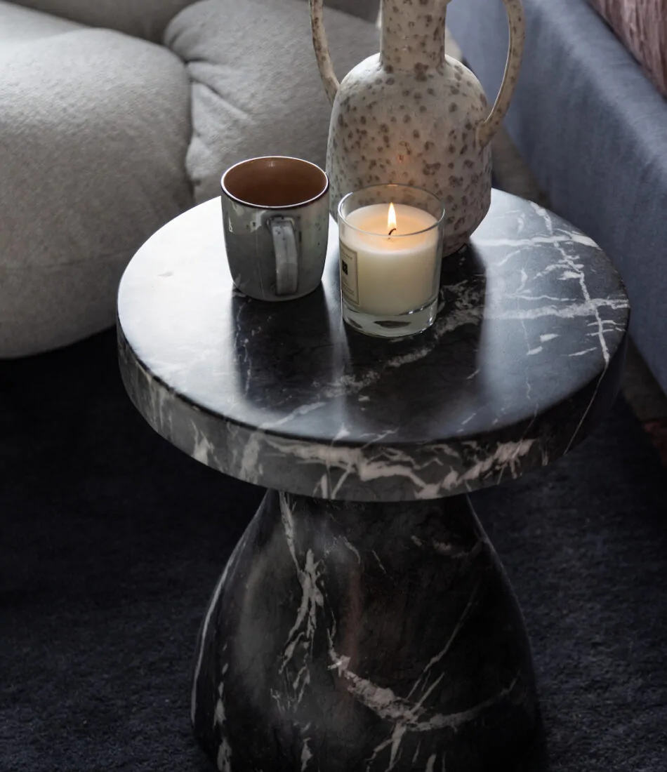 A round black marble table holds a decorative pitcher, a ceramic mug, and a lit candle, set on a dark rug beside a light-colored, plush armchair.