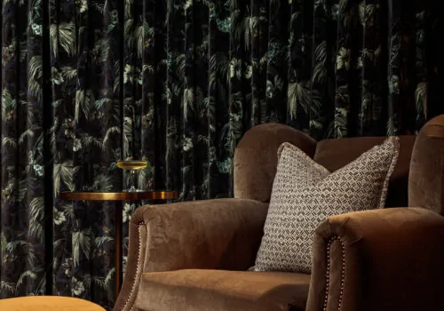 Home Cinema Room - Cosy muted brown armchair with burnt orange velvet fluted footstool, curved curtain track with dark floral curtains