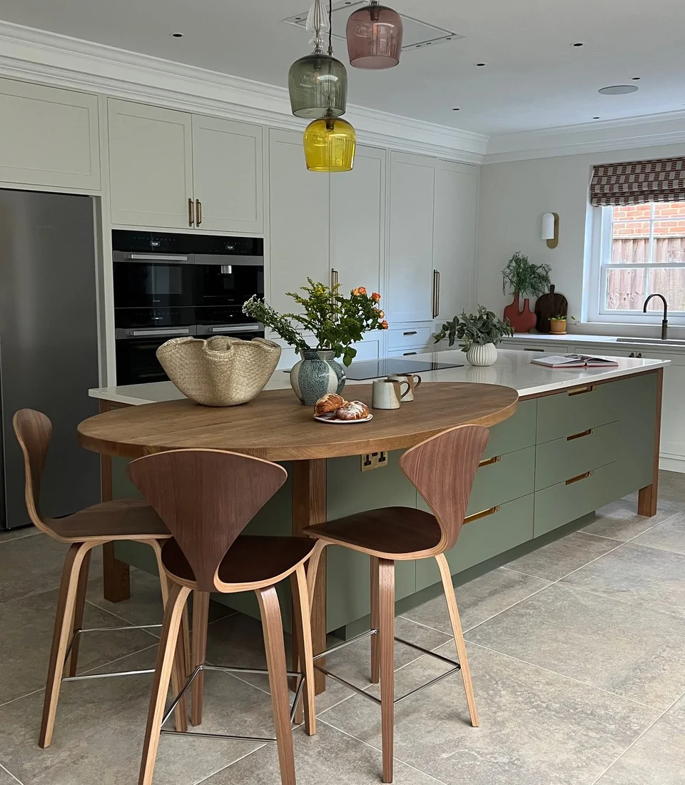 A round wooden table with two modern chairs holds a vase of flowers and a plate of pastries. The kitchen features light cabinets, stylish pendant lights, and a bright window.