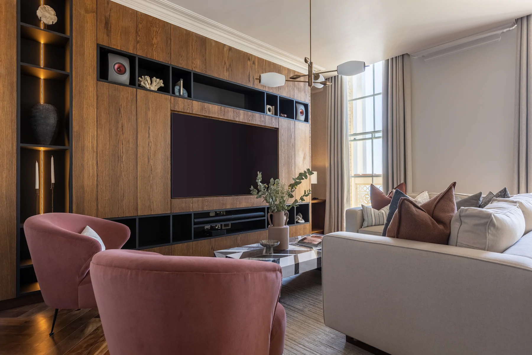 A modern living room features two pink chairs and a light-colored sofa, with a wooden entertainment unit holding a television and decorative items. Sunlight streams through large windows.