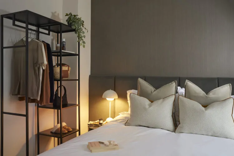 A stylish bedroom features a black metal shelving unit with clothes and accessories, a cozy bed with plush pillows, and a soft bedside lamp casting warm light, enhancing a serene atmosphere.