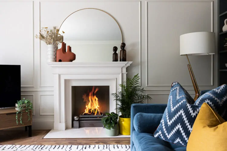 A cosy living room features a white marble fireplace with a dancing flame, surrounded by decorative vases and a mirror. A blue sofa with patterned cushions sits nearby, with greenery enhancing the warmth.