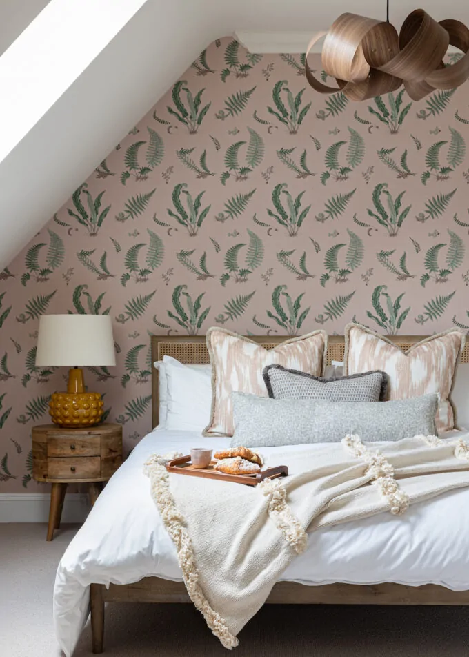 A cosy bedroom features a bed with decorative pillows and a blanket, a wooden bedside table with a lamp, and a tray of pastries. The walls are adorned with botanical-patterned wallpaper.