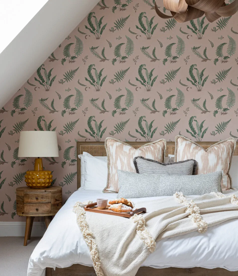 A cosy bedroom features a bed with decorative pillows and a blanket, a wooden bedside table with a lamp, and a tray of pastries. The walls are adorned with botanical-patterned wallpaper.