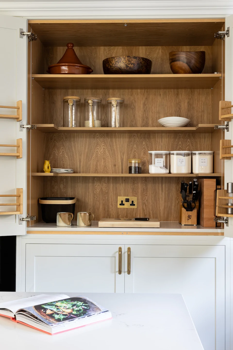 A kitchen cabinet displays various items including a terracotta pot, wooden bowls, glass jars, and coffee containers. A countertop holds a cookbook and cutting board, with utensils neatly arranged.