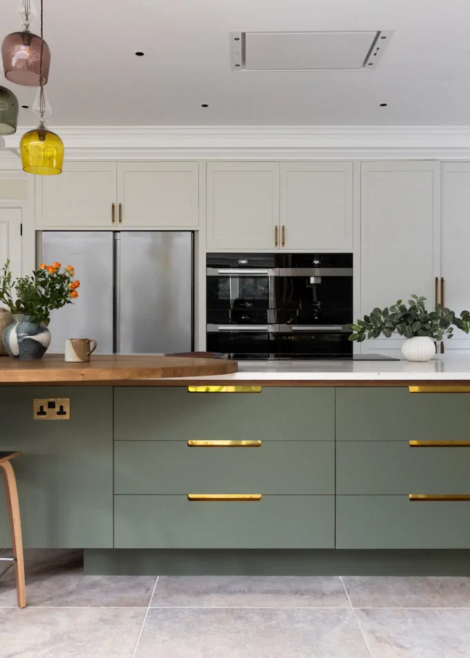 A green kitchen island with wooden countertops displays a vase of flowers and baked goods. A person in motion passes by, while colorful pendant lights hang above. The context is a modern kitchen setting.