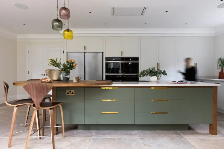 A green kitchen island with wooden countertops displays a vase of flowers and baked goods. A person in motion passes by, while colorful pendant lights hang above. The context is a modern kitchen setting.