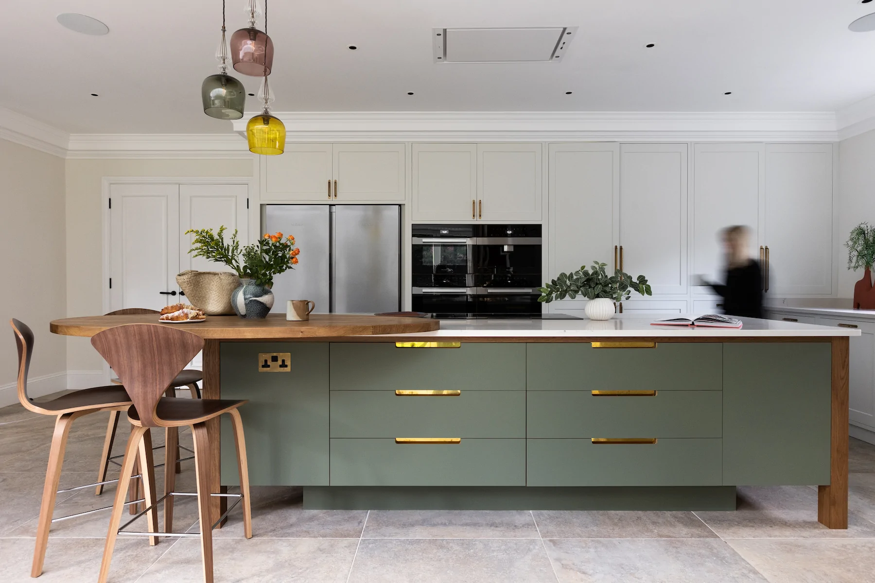 A green kitchen island with wooden countertops displays a vase of flowers and baked goods. A person in motion passes by, while colorful pendant lights hang above. The context is a modern kitchen setting.