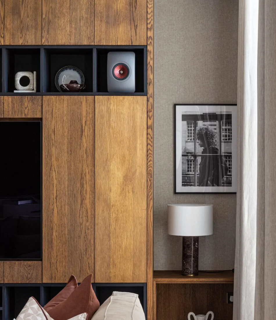 A wooden wall unit displays decor items, while a cozy sofa with patterned pillows sits in front. A lamp and a decorative vase are placed on a side table nearby. A framed black-and-white photo hangs on the wall.