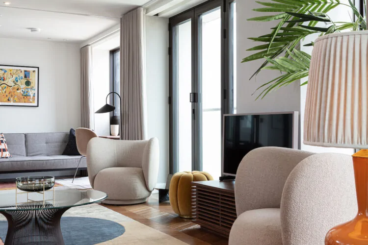 A cozy living room features soft, rounded chairs and a round coffee table atop a multicolored rug. Natural light streams through large windows, illuminating a modern TV and decorative plant.