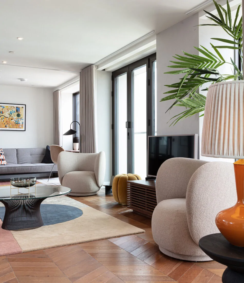 A cozy living room features soft, rounded chairs and a round coffee table atop a multicolored rug. Natural light streams through large windows, illuminating a modern TV and decorative plant.