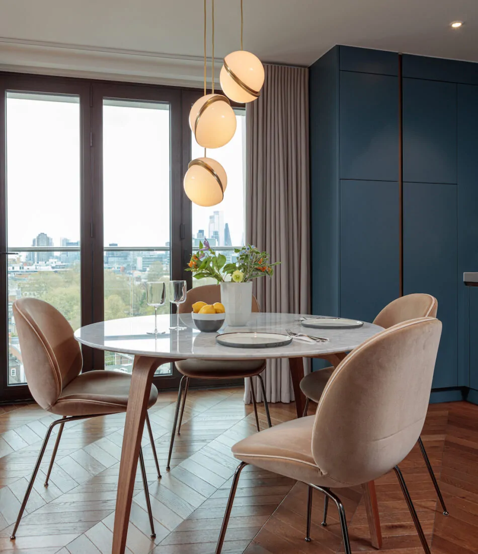 A round marble dining table with elegant chairs is set with glassware and plates. A bouquet of flowers and lemons adorn the table, illuminated by hanging pendant lights, against a scenic urban view through large windows.