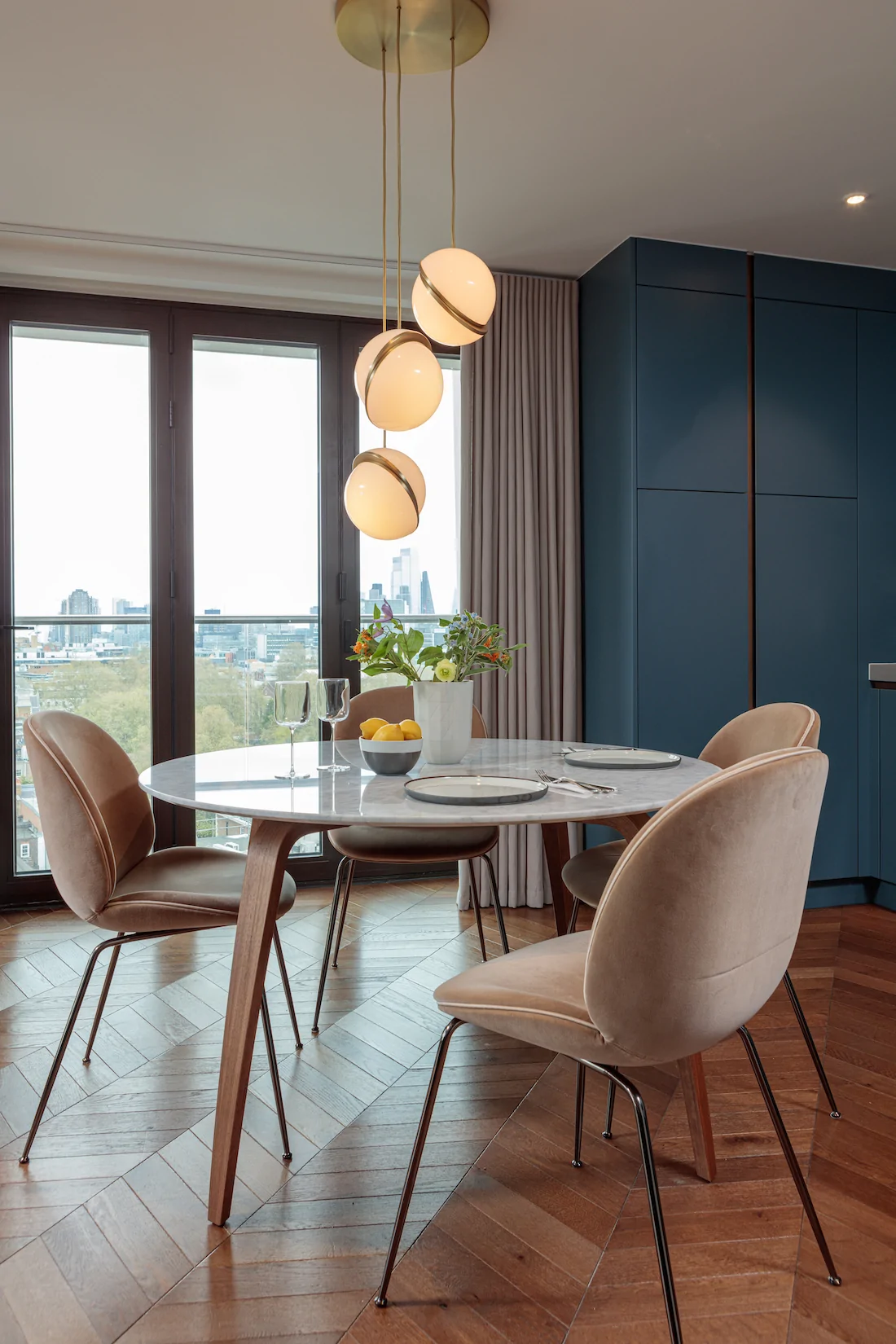 A round marble dining table with elegant chairs is set with glassware and plates. A bouquet of flowers and lemons adorn the table, illuminated by hanging pendant lights, against a scenic urban view through large windows.