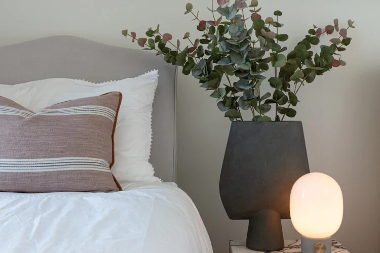 A plush bed with decorative pillows sits beside a marble side table. An abstract art piece adorns the wall, while a modern lamp and greenery complete the cozy bedroom scene.