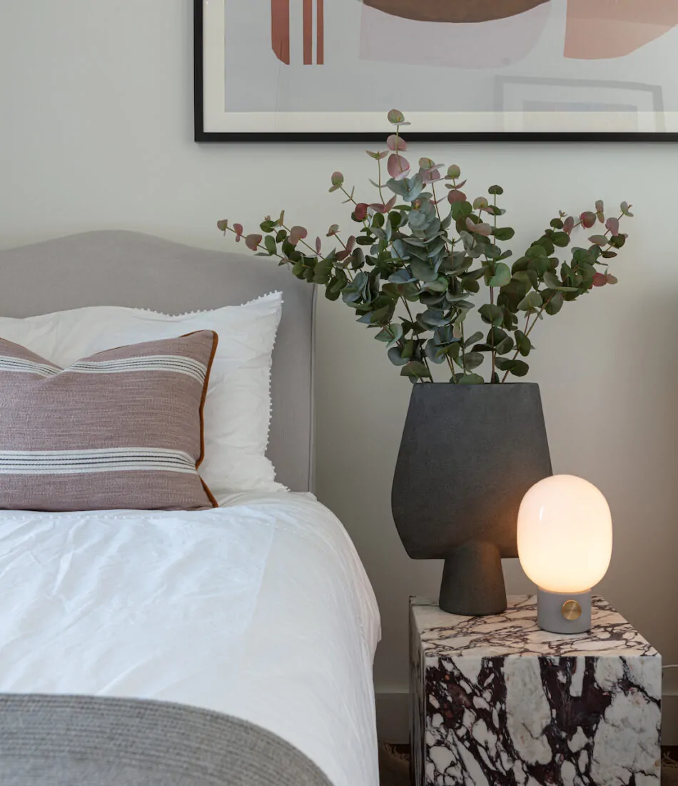 A plush bed with decorative pillows sits beside a marble side table. An abstract art piece adorns the wall, while a modern lamp and greenery complete the cozy bedroom scene.